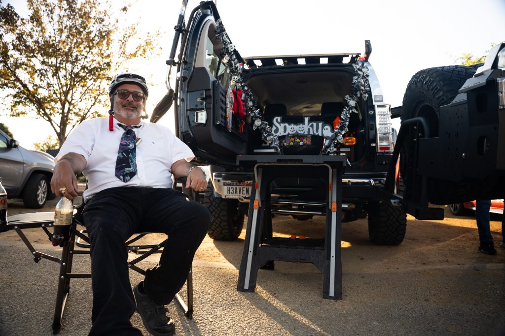 Costumed man in a shirt and tie seated beside a Halloween-decorated SUV at Trunk or Treat