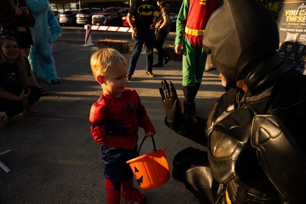 Young boy in a Spider-Man costume holding a pumpkin bucket and meeting Batman at Trunk or Treat