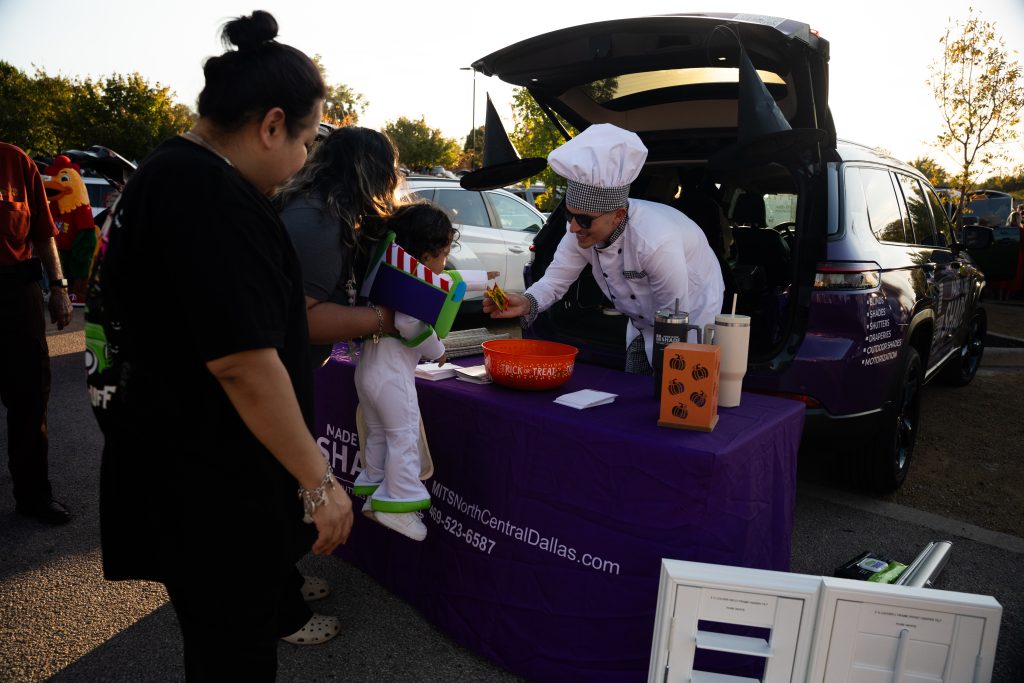 Vendor in a chef's hat handing treats to a child at a purple-draped trunk during Trunk or Treat