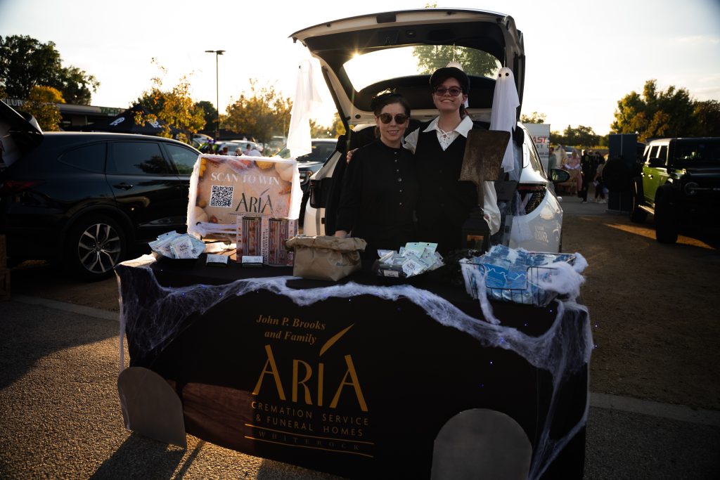 Two vendors behind a Halloween-decorated ARIA table and open trunk at Trunk or Treat, Lake Ridge Village