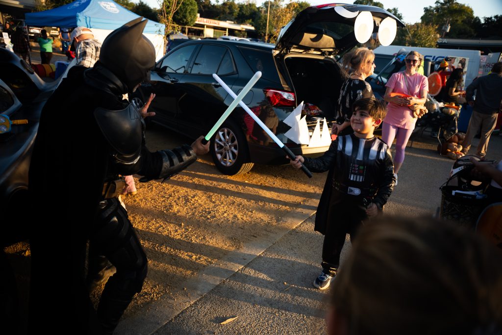 Child in costume crossing lightsabers with an armored character among decorated trunks at Trunk or Treat
